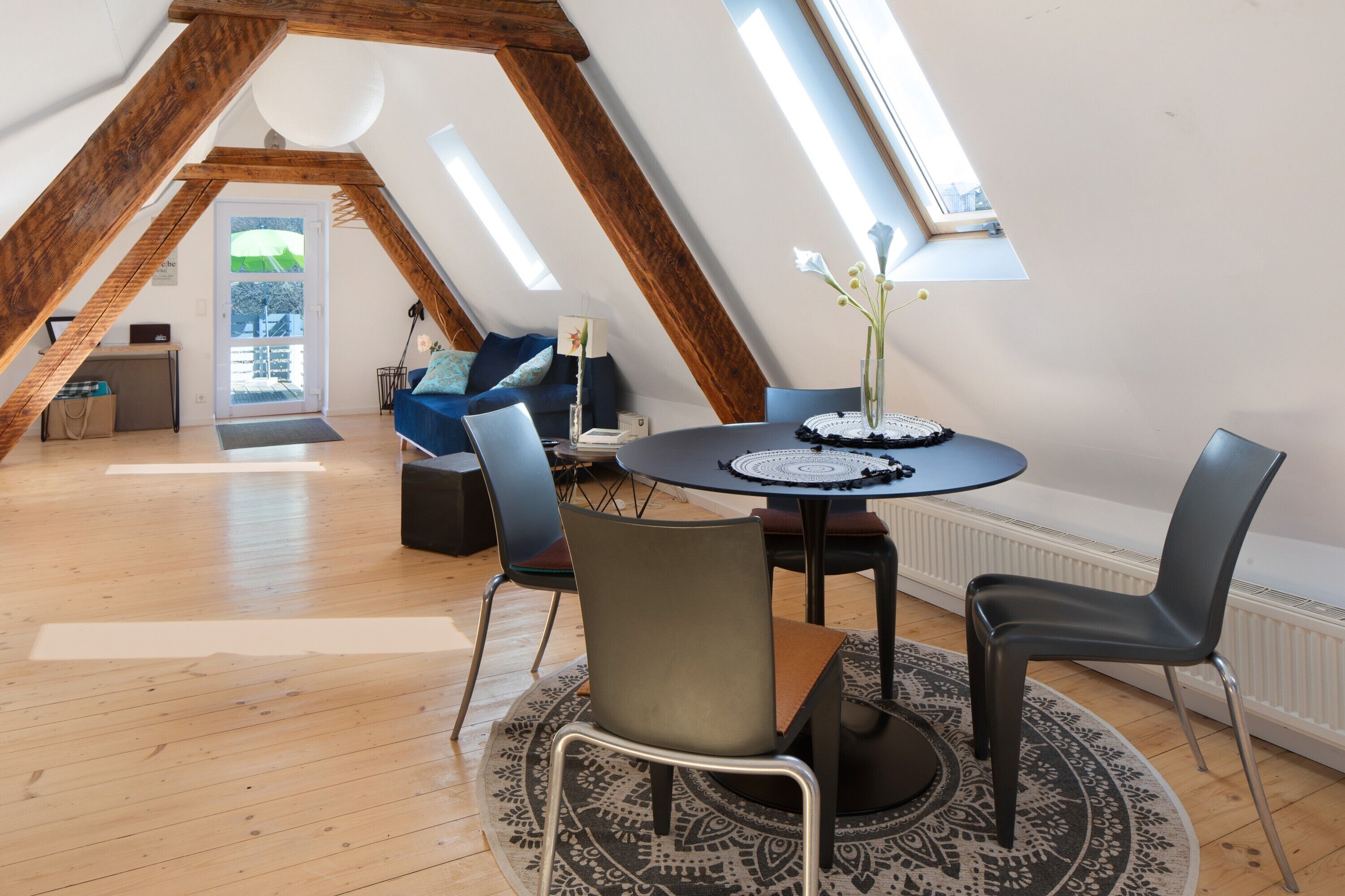 Una stanza mansardata con soffitto in legno, un tavolo rotondo con sedie, un vaso con fiori e una vista attraverso una porta.