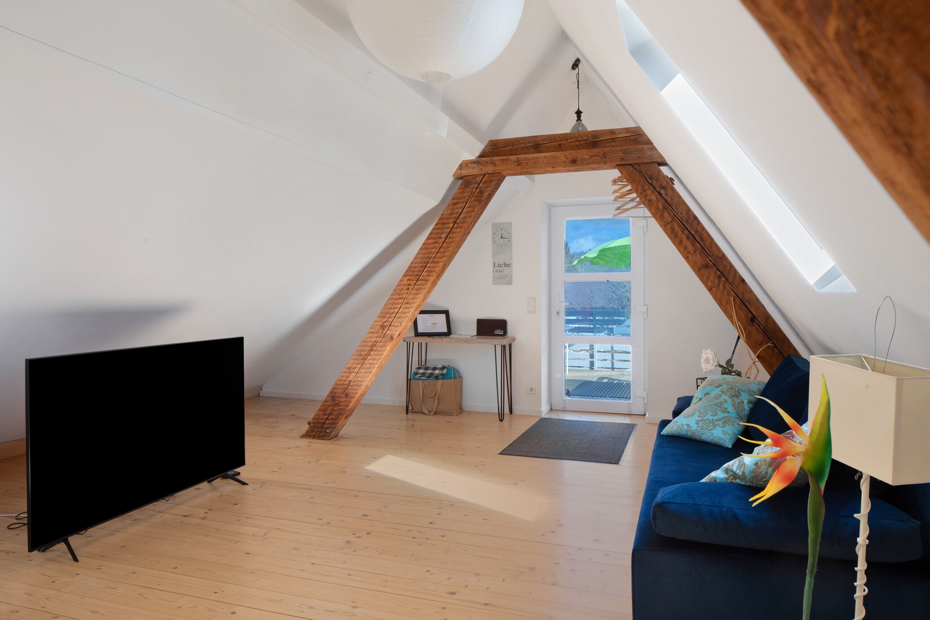 A room with a sloped ceiling, a television, a sofa, a lamp, and a window with a view.