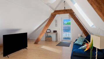 A room with a sloped ceiling, a television, a sofa, a lamp, and a window with a view.