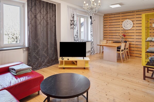 Ein Wohnzimmer mit rotem Sofa, einem schwarzen Tisch, einem Fernseher auf einem Holzboden, einer Kristallleuchter, einer Uhr und einem Essbereich mit Stühlen und einem gelben Schrank.