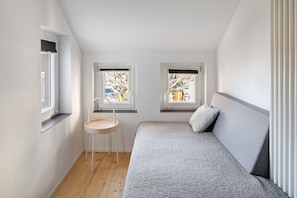 A bedroom with a bed, two windows, a chair, and a small table.