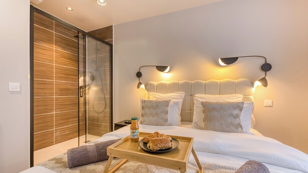 A hotel room with a bed, bedside lamps, a tray with croissants, a bottle of juice, and a shower area.