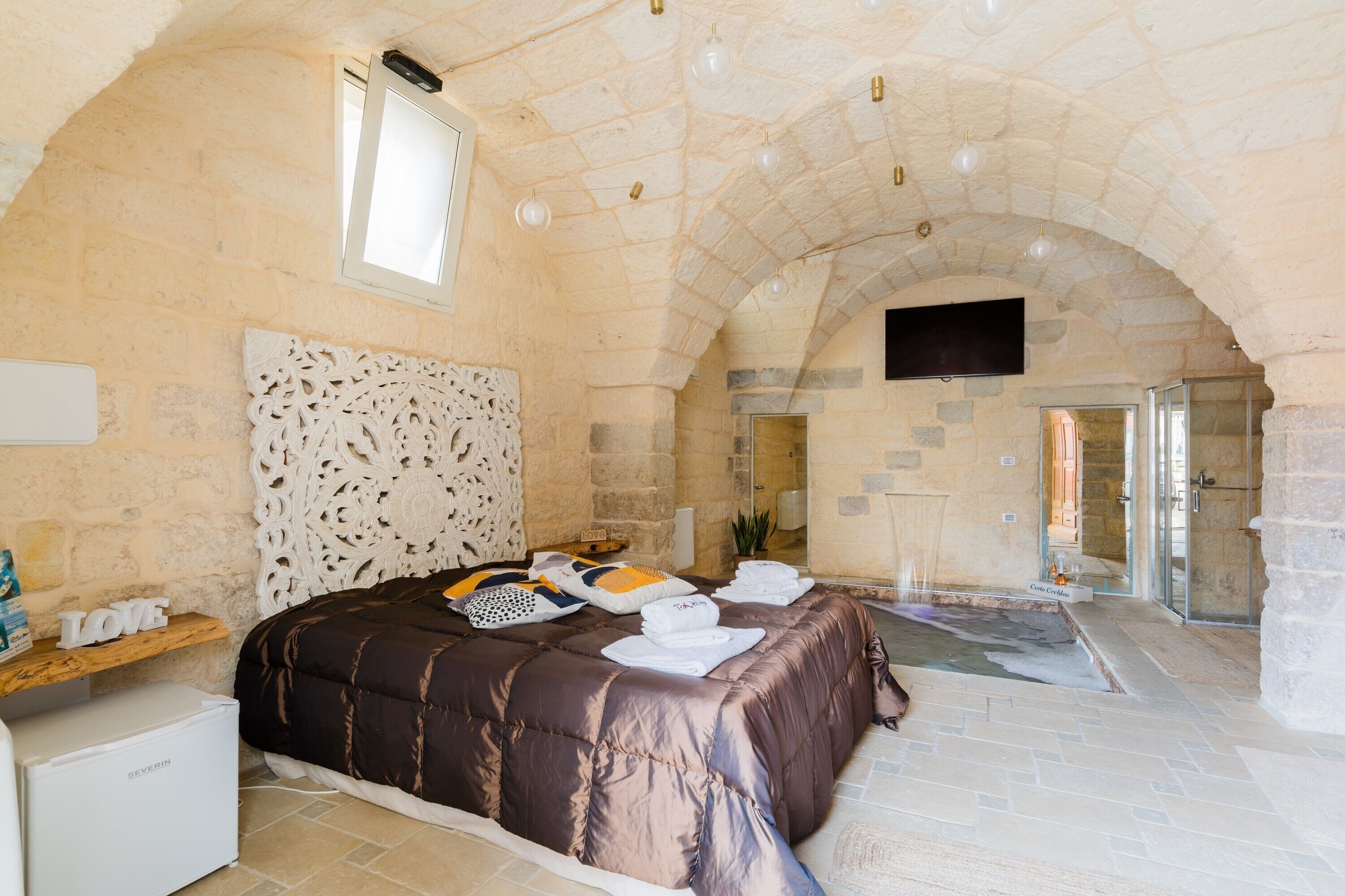 Un dormitorio con techo abovedado de piedra, cama con colcha marrón, un refrigerador pequeño y zona de ducha.
