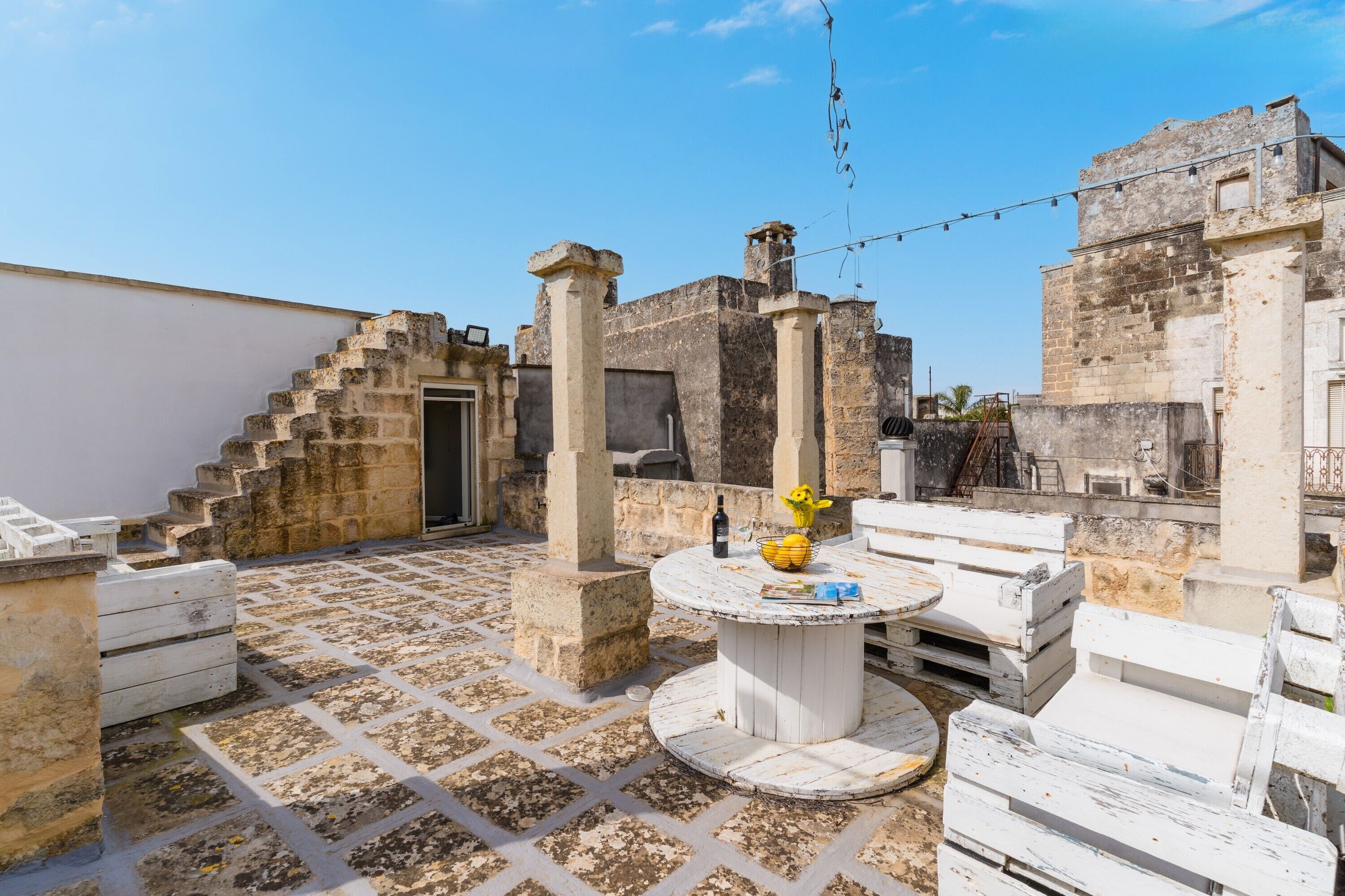 A courtyard with stone flooring, a round table with a bottle and a yellow bowl, and a staircase leading to an upper level.