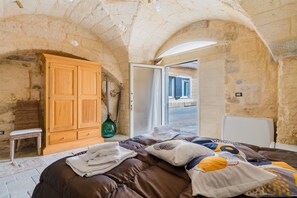 Une chambre avec des murs en pierre, une armoire en bois, un lit recouvert d’une couverture marron et une chaise en osier.