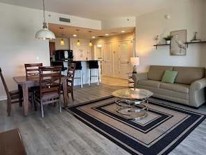 A modern living room with a dining area, a sofa, and a kitchen in the background. - 6th Floor Cozy Beach Retreat at Origin at Seahaven  - Daily Free Activities! (Panama City Beach)