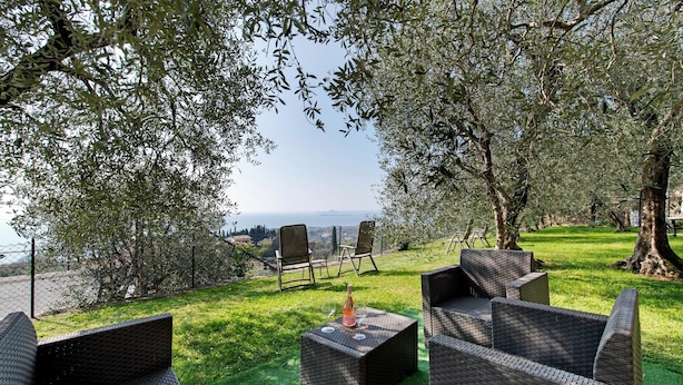 An outdoor seating area with wicker furniture, a bottle, and a glass, surrounded by trees and a view of the sea.