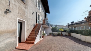 A building with a staircase leading to a door, a paved courtyard, and a hedge.