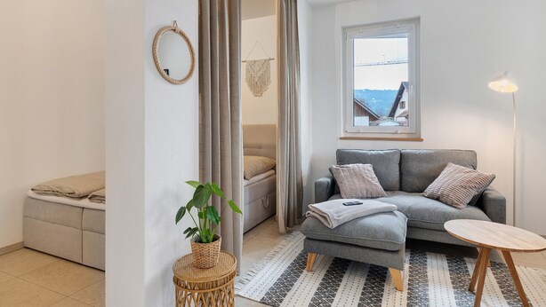 A modern living room with a grey sofa, a small round table, a potted plant, and a view of a bridge through a window. - Apartment Bodensee Lindau – Modernes Apartment mit Terrasse und Bergblick (Lindau (Bodensee))
