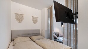 A bedroom with a bed, two macramé wall hangings, a television, and a view of a living area through a curtain. - Apartment Bodensee Lindau – Modernes Apartment mit Terrasse und Bergblick (Lindau (Bodensee))
