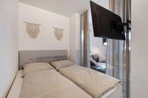 Une chambre à coucher avec un lit, deux décorations murales en macramé, un téléviseur et une vue sur le salon à travers un rideau.