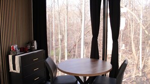Una habitación con una mesa redonda de madera y dos sillas, un aparador negro con cajones, y vistas a árboles sin hojas a través de una ventana.