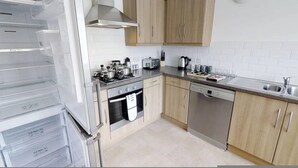 A modern kitchen with wooden cabinets, a stainless steel dishwasher, a stove, and a refrigerator with transparent drawers. - Cole Green Lane Executive Stay- Free Parking (Welwyn Garden City)