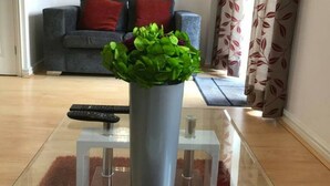 A living room with a grey sofa, a potted plant, a remote control, and a glass coffee table. - Cole Green Lane Executive Stay- Free Parking (Welwyn Garden City)