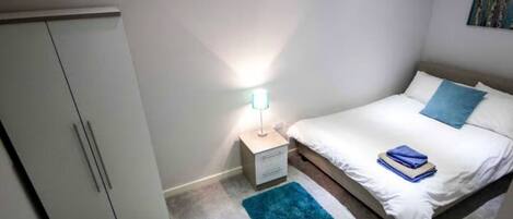 A bedroom with a bed, a nightstand, a lamp, a wardrobe, and a blue rug.