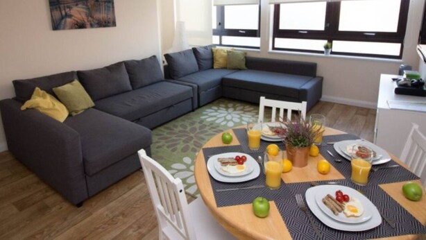 A modern living room with a sectional sofa, a dining table set for breakfast, and large windows with blinds. - Castle Point 6 Apartment - Premier Lodge (Southampton)