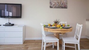 A dining area with a round table, white chairs, a flat-screen TV mounted on the wall, and a floral painting above the table. - Castle Point 6 Apartment - Premier Lodge (Southampton)