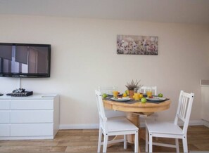A dining area with a round table, white chairs, a flat-screen TV mounted on the wall, and a floral painting above the table. - Castle Point 6 Apartment - Premier Lodge (Southampton)