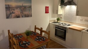A compact kitchen with white cabinets, a stove, and a sink. There's a wooden dining table with chairs and a placemat featuring a cartoon character. A painting of a beach scene hangs on the wall. - Stylish Accommodation in Southampton 10 (Southampton)