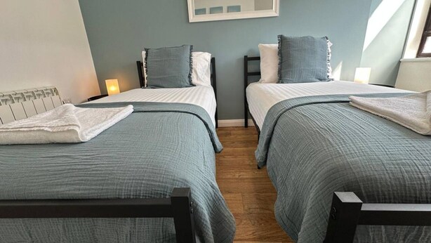 A room with two beds, each with a grey bedspread and pillows, a white radiator, a mirror on the wall, and a window with natural light. - Stylish Accommodation in Southampton 10 (Southampton)
