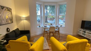 A living room with a sofa, a dining table, chairs, a TV, and a window with a view of the street. - Charming Bedford Apartment 1 Near Bedford Park (Bedford)