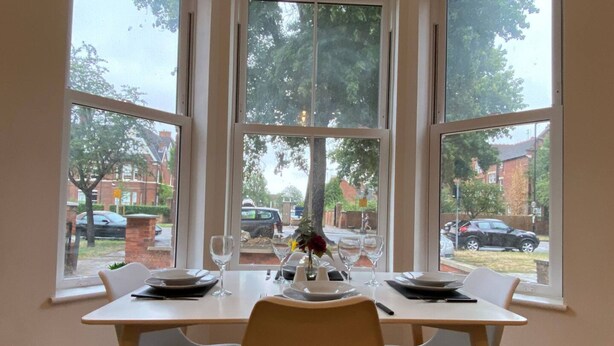 A dining table set for two with a vase of flowers, large windows with a view of a residential area. - Charming Bedford Apartment 1 Near Bedford Park (Bedford)