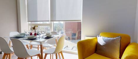 Un comedor con una mesa lista para cuatro personas, sillas blancas, un sillón amarillo y una ventana con persianas.