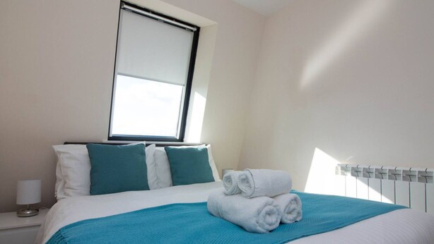 A hotel room with a bed, a window, a nightstand, and a rolled towel on the bed. - Premiere Serviced Accommodation - 17 (Southampton)