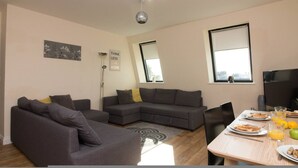A modern living room with a grey sectional sofa, a dining table with breakfast items, and large windows. - Premiere Serviced Accommodation - 17 (Southampton)