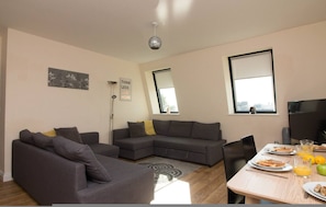 A modern living room with a grey sectional sofa, a dining table with breakfast items, and large windows. - Premiere Serviced Accommodation - 17 (Southampton)
