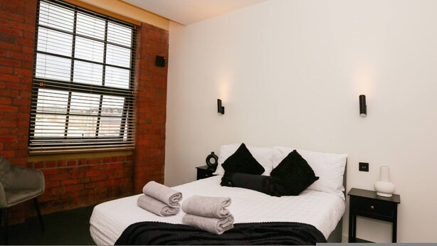 A hotel room with a brick wall, a large window with blinds, a bed with white bedding and black pillows, two bedside tables, and a chair. - Nichols Building City Escape Spacious 3BR Sheffield (Sheffield)