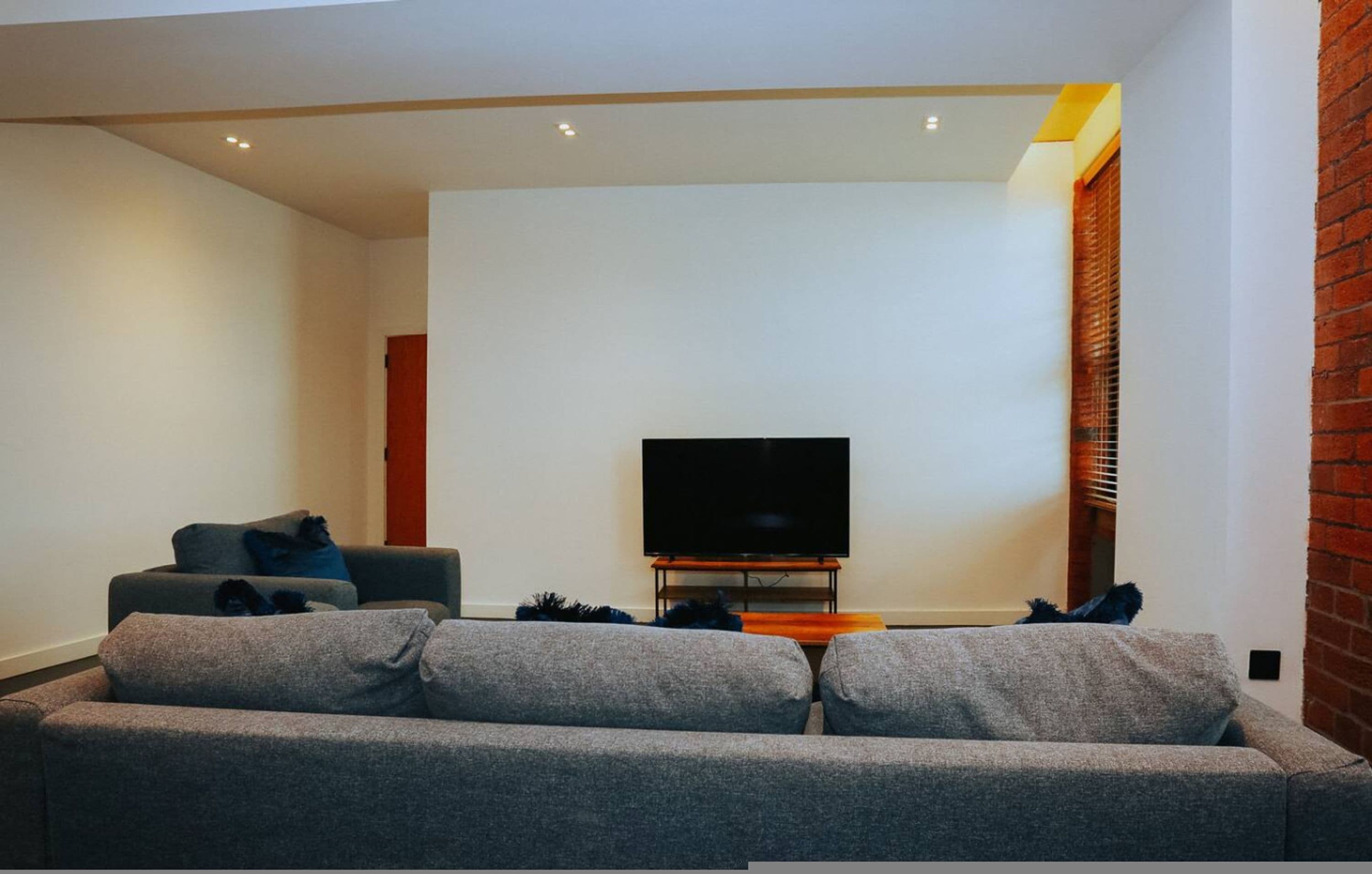Un salon avec un canapé gris, un téléviseur à écran plat posé sur un meuble en bois et un mur en briques.
