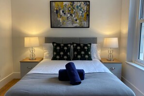 A neatly made bed with white and dark green pillows, two bedside lamps, and a blue towel on the bed. - Stylish Bedford Town Centre Apartment 5 Sleeps 6 Fast Wi-fi (Bedford)