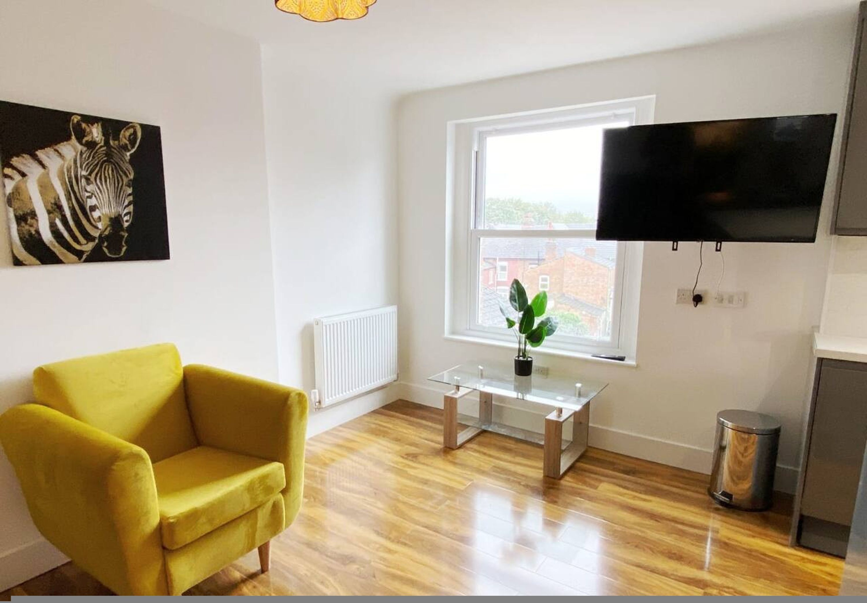 A room with a yellow armchair, a glass table with a plant, a mounted TV, and a window with a view of buildings.