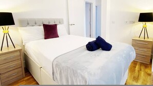 A neatly made bed with white bedding, a dark red pillow, and a folded blue towel. There are two bedside tables with lamps, one on each side of the bed. - Stylish Bedford Town Centre Apartment 5 Sleeps 6 Fast Wi-fi (Bedford)