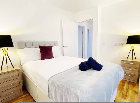 A neatly made bed with white bedding, a dark red pillow, and a folded blue towel. There are two bedside tables with lamps, one on each side of the bed. - Stylish Bedford Town Centre Apartment 5 Sleeps 6 Fast Wi-fi (Bedford)
