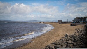 Una playa con costa rocosa, un muelle que se adentra en el agua y edificios a lo largo de la costa.