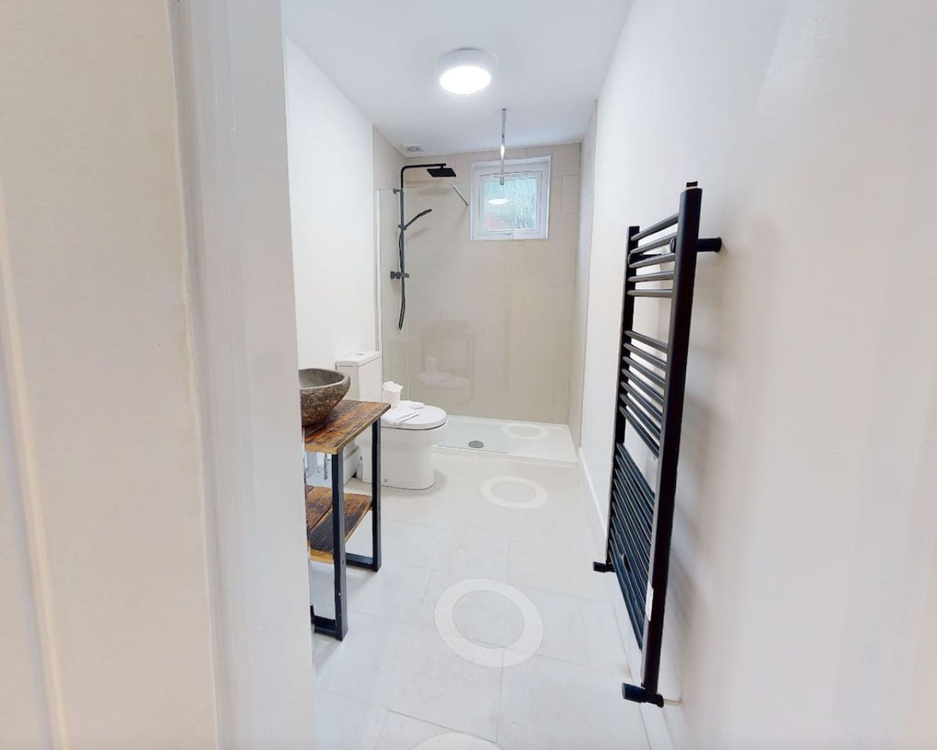 A narrow hallway leading to a bathroom with a toilet, shower, and a wooden table with a bowl.