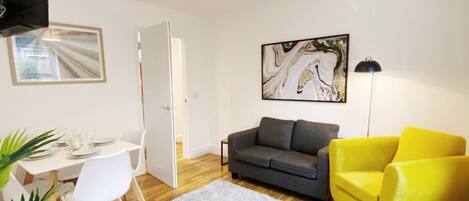 A living room with a grey sofa, a yellow armchair, a white dining table with chairs, a white rug, and a painting on the wall.