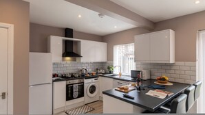 A modern kitchen with white cabinetry, a black range hood, a built-in oven, a washing machine, and a dining area with a table and chairs. - Buckingham House Modern Coastal 3BR Stay (Morecambe)