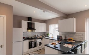 Una cocina moderna con armarios blancos, una campana extractora negra, horno empotrado, lavadora y comedor con mesa y sillas.