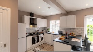 A modern kitchen with white cabinetry, a built-in oven, a washing machine, and a large window providing natural light. - Buckingham House Modern Coastal 3BR Stay (Morecambe)
