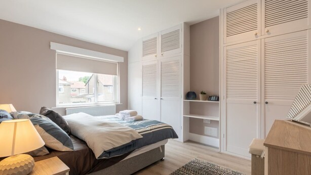 A bedroom with a bed, bedside lamp, window, and built-in wardrobe with louvered doors. - Buckingham House Modern Coastal 3BR Stay (Morecambe)