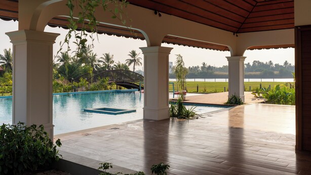 A covered outdoor area with a pool, columns, and a view of a bridge and palm trees. - VIHARA By CMK (Kanayannur)