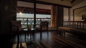 A room with a wooden floor, a sofa, two chairs, a small table with a plant, and a view of a body of water through large windows. - VIHARA By CMK (Kanayannur)