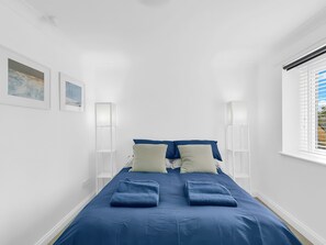 A narrow, white-walled room with a bed centered, flanked by two bedside tables with lamps, a window with blinds, and two framed pictures on the wall. - Central Sherborne (Sherborne)