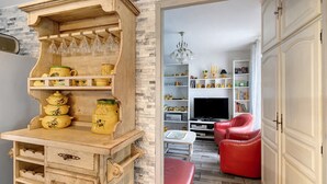 Eine Küche mit Holztisch, Weingläsern und gelben Gläsern. Im Wohnbereich steht ein roter Sessel und ein Fernseher.