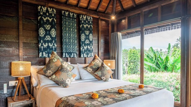 A wooden-framed room with a bed, a nightstand, and a window with a view of greenery. - Airlangga Dsawah by Balihora, Ubud Vill (Ubud)