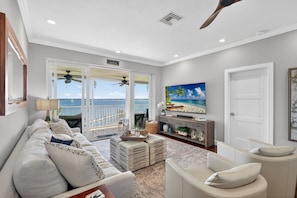 A living room with a sofa, ottoman, TV, and a view of the ocean through glass doors. - Warm Florida Sun with Spectacular River Views in Peaceful Active Community (Stuart)