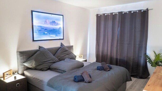 A bedroom with a bed, pillows, a nightstand, a wall-mounted TV, and a window with curtains. - Apartment Fioranza Comfortable stylish apartment for 6 people (Mülheim an der Ruhr)